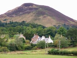 A small cluster of houses sit among trees at the base of a tall bare hill in the background.