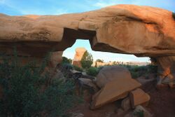 Mano Arch—much thicker than Metate Arch—with several boulders lying under it and a large notch missing from the bottom of the arch. A distant hoodoo appears framed inside the arch's notch.