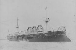 A large, black ship with three short smoke stacks has two small white boats tied alongside.