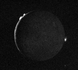 The thin crescent (open to the right) of the full disk of a planetary body with two bright clouds along the upper left edge of the object and another along the right edge.