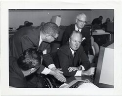 Photograph of four men in suits sitting in front of a computer.