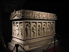Three quarters view of a decorated marble sarcophagus in the shape of a Greek temple.