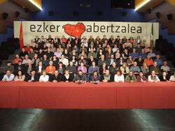 A table with seven steps of people with a banner ezker abertzalea in the background and the flags of Navarre on the left and the Basque Country on the right.