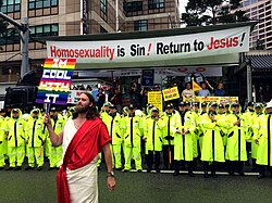 A man with long hair and a beard, dressed as Jesus, holds up a sign reading "I'm Cool With It" atop a rainbow background. He is standing in front of a group of protesters holding anti-LGBT signs on the back of a semi truck, underneath a banner reading, "Homosexuality is Sin! Return to Jesus!". A line of police stand in between, guarding the protesters.