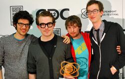 Four males from the development team stand with an atom-like trophy with a GDC backdrop. From left to right, Vreeland has long, messy hair, rectangular glasses, a TV static-like sweater, and is unshaven. His arm is around Fish, whose brown hair is combed back. He wears thick black glasses frames and a black shirt underneath a gray cardigan. McCartin has brownish-red hair and a goatee and wears a Fez logo T-shirt underneath a red zippered hoodie. Bédard is a head taller than the bunch, and has short, brown hair, rectangular glasses, and is unshaven. He wears a black and white checkered dress shirt, a black tie, and a zippered, black hoodie.