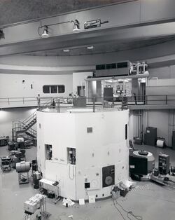 The black-and-white photograph is of a large room that contains a lot of electronic equipment. The lower half of the image contains a cylindrical white container that is a nuclear reactor. There is a walkway at the top of the reactor, which leads back to a control room where two men are sitting.