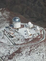 Looking down from the sky at a series of buildings and a large domed telescope building that dwarfs the others. An access road encircles the complex.
