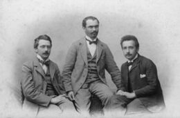 Three young men in suits with high white collars and bow ties, sitting.