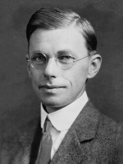 Head and shoulders of smiling man in suit and tie with round dark-rimmed glasses. This is a detail from the picture below.