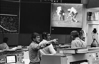 A control room; visible on a large screen are two astronauts walking on the Moon