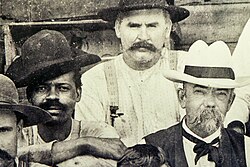Three men sit for a black and white photo. On the right is a caucasian man with a large mustache and trimmed beard, wearing a white hat with a black hatband. This is Jack Daniel, the founder of Jack Daniel's Tennessee Whiskey. On the left is an African-American man with a smaller black mustache wearing a simple black hat. This man is theorized to be Nathan "Nearest" Green or possibly one of his sons. Behind the Daniel and Green is an unknown caucasian man.