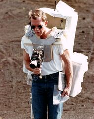 Man around age 40 with sunglasses and a large backpack takes a photograph with a camera mounted on his chest