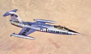 Jet fighter in metallic scheme with T-tail and short wings flying above desert and black constructions