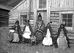 A black and white photo of six indigenous people in front of a log cabin.