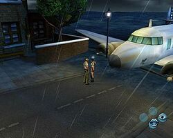 A screenshot of two people, a man and a woman, standing in front of an aeroplane; there are four circles in the lower right of the screen in a trapezoidal pattern, with a mouth in the lowest and an eye in the right-hand one