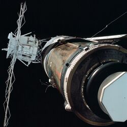 An image of Skylab. The left side of the frame is dominated by a communications array, painted white with a cylindrical satellite dish on top. On they right is a brown-grey cylinder, which is the main station. No solar arrays are visible.