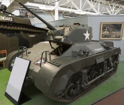 A squat dark green tank in a museum, placed on a large green felt mat. There is a description board in front of it, and a grey divided wall to its rear. The tank's gun barrel is pointing towards the ceiling and its left-hand caterpillar tracks are visible, as well as several large handles on its hull.