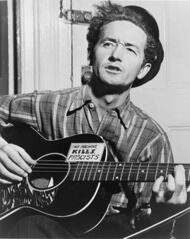 A black-and-white photograph taken indoors of Woody Guthrie wearing pinstripe trousers, a tartan shirt with the top button undone, and a cap. He sits playing a six-string acoustic guitar, which is supported on one knee, and he appears to be singing. 'This Machine Kills Fascists' is written in all capital letters on a rectangular sticker, which is fixed onto the guitar.