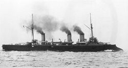 A large warship with two large guns steams at high speed, with thick black smoke pouring from its three funnels.