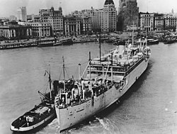 USS Chaumont, a large light-coloured transport vessel with a smaller vessel alongside, against a background of shorefront buildings and docked ships