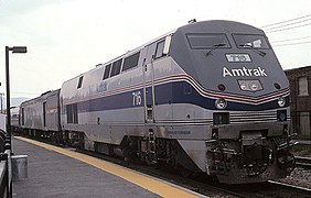 A light gray diesel locomotive with a dark gray top stripe, a middle blue stripe, and two thinner red stripes on the side. The stripes narrow and angle downwards on the front.