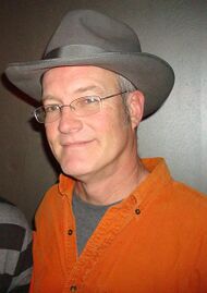 A bespectacled man, wearing an orange shirt, a gray t-shirt and a fedora, smiles at the camera.