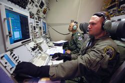Inside military aircraft. Two personnel manning communications consoles with wide displays.