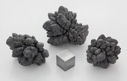 A small gray metal cube surrounded by three gray metal nuggets in front of a light gray background