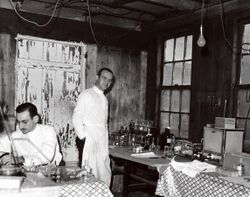 Two scientists in white lab coats, one seated and one standing, in a small building with peeling paint, standing next to a table with scientific glassware