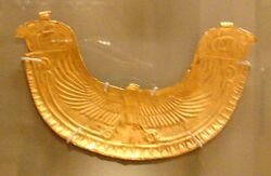 Image of a gold pectoral in the shape of an eagle spreadingh its wings. The Wings end in two Horus heads at the tip of each of the wings