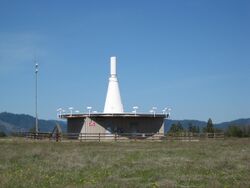A thin up-pointing white cone with a cylindrical tip, a few meters tall, sits atop an elevated concrete platform, seen against clear blue sky; it is surrounded by a circle of about a dozen small picket-like vertical antennas