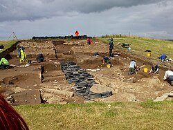 A large rectangular trench in which a group of people are digging or recording archaeological features.