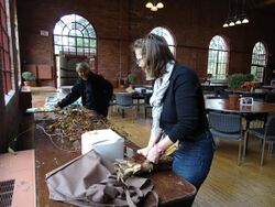 Two people stand at a long table preparing autumnal items for an event.