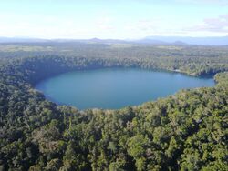 Aerial view of Lake Eacham, surrounded by dense tropical rainforest, with the agricultural land use of the Atherton Tablelnds in the background.