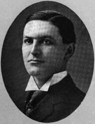 A black and white photograph of a young white man with a short haircut and his shirt collar turned slightly up over his tie