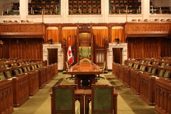 A photograph of a lit room in which there are many brown-and-green chairs facing in various directions as well as a brown table