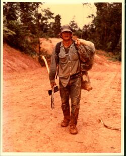 man standing on dirt road with muddy pants and shoes carrying tools and a bag