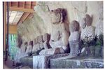 Photograph of a row of ca. ten seated stone statues in front of a rock. Three-quarter view. One of the statues is about twice as large as the others.
