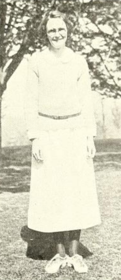 A young white woman, standing outdoors and smiling, wearing glasses and a long light-colored dress with long sleeves and a thin belt