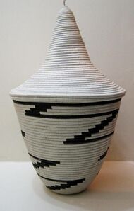 Photograph depicting a bowl shaped off-white woven basket with tall conical lid and black zigzag pattern
