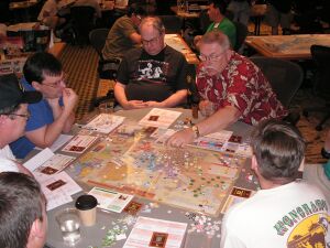 Six men sit around a table, studying the contents atop it. On the table are a map, many small cardboard chits (counters), and cards. One man has his left hand hovering above the map, finger pointing at a stack of counters.