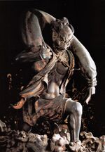 Fūjin. Three-quarter view of a statue. His left leg is bent as if climbing stairs and he is carrying a long bag-shaped object which goes from one shoulder to the other around the back of his head. Black and white photograph.