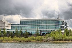 A modern three-storey office building with reflective greenish glass windows underneath a cloudy sky along the lakeshore, seen from the opposite side
