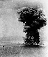 A view of the ocean stretching to the horizon with the silhouette of a distant small warship visible to the left. To the right an enormous mushroom cloud rises high into the sky.