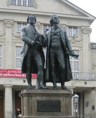 Photograph of a large bronze statue of two men standing hand-in-hand, side by side and facing forward. The statue is on a stone pedestal, which has a plaque that reads "Dem Dichterpaar/Goethe und Schiller/das Vaterland".