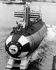 Black-and-white photo of submarine being slid into water during launching. Dozens of people are standing on top of it.