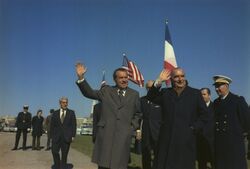 Two men in suits stand to the right, with uniformed military officers nearby. Both men are waving and smiling.