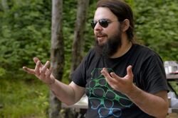 McGrath gesticulates with both hands while looking to the left. He wears aviator sunglasses, a black T-shirt with a sunglasses graphic, and has a long, brown beard. He is in an outdoors, camping setting.