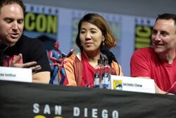Left to right, a caucasian male with short brown hair, an Asian woman with medium length brown hair and a caucasian male with short black hair sit behind a table. A small statue of a punk version of Spider-Man sits on the table in front of them.