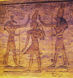 Relief of a man with an elaborate crown between two figures who gesture toward the crown. The figure on the left has the head of an animal with square ears and a long nose, while the one on the right has a falcon's head.